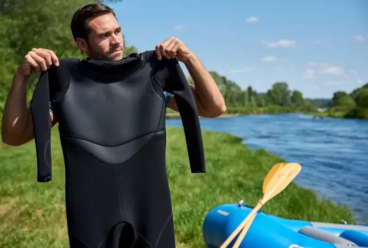 Person considering wearing a wetsuit beside rafting gear near a river under clear daylight, illustrating the guide's topic.