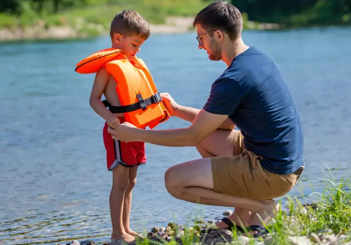 Parent helping child adjust a PFD near calm water under clear daylight, symbolizing assessing family readiness for rafting.
