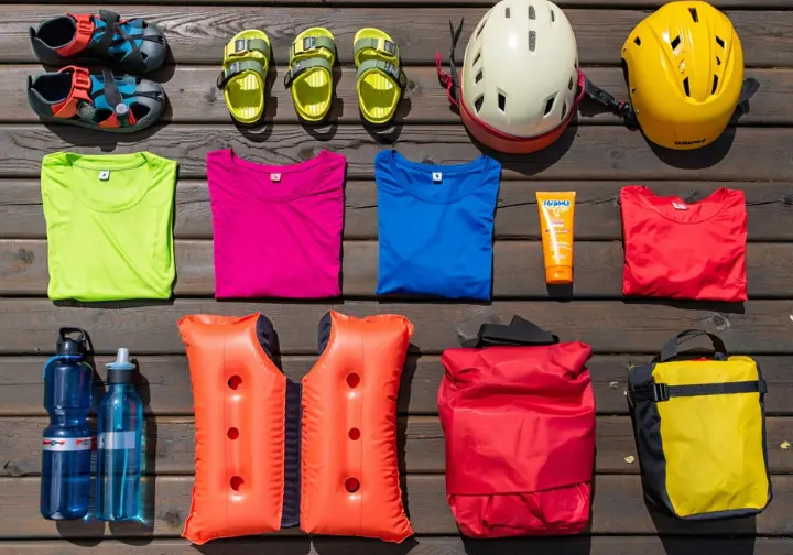 Neatly arranged essential family rafting gear (synthetics, PFDs, sunscreen) in bright daylight, illustrating smart packing.