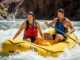 PVC vs Hypalon Rafts: A Guide to River Class & Material A full-body shot of a young man and woman in swimwear and life jackets paddling an inflatable raft through whitewater on a sunny day.