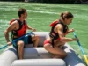 Master the Rafting Draw Stroke: From Eddies to Ferries A fit young woman in a bikini and life jacket executes a draw stroke to guide a raft, while a man in boardshorts sits in the front on a sunny river.