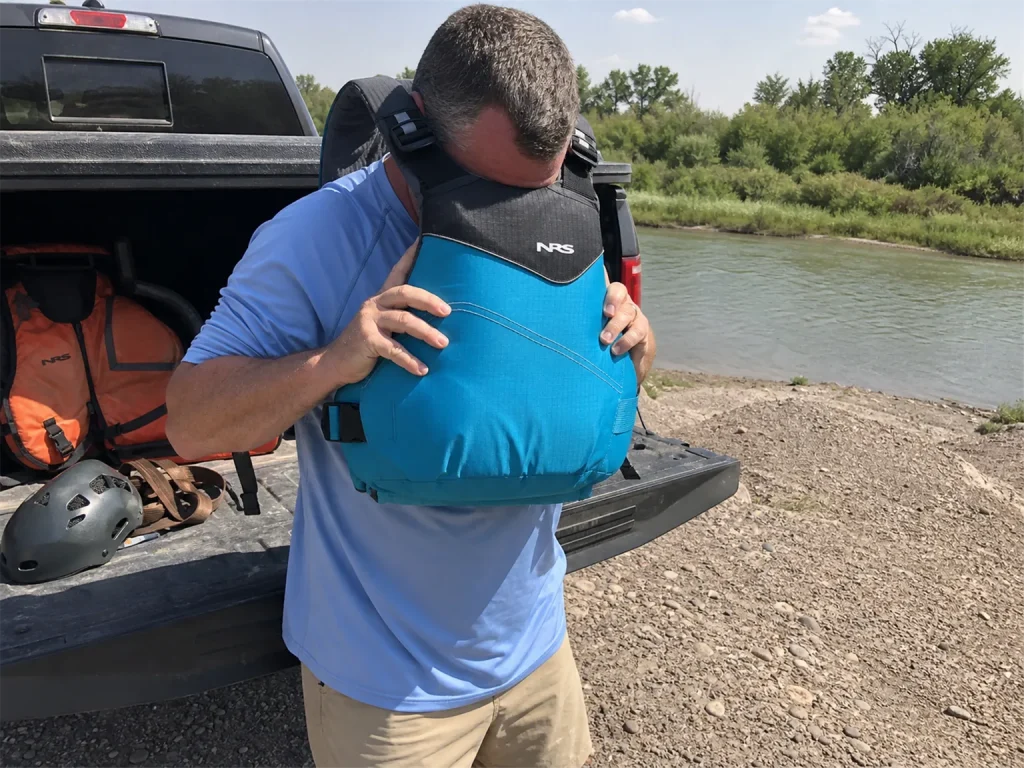 Boater pulling an NRS Vapor rafting PFD over his head at a truck tailgate, old orange boat vest in the bed