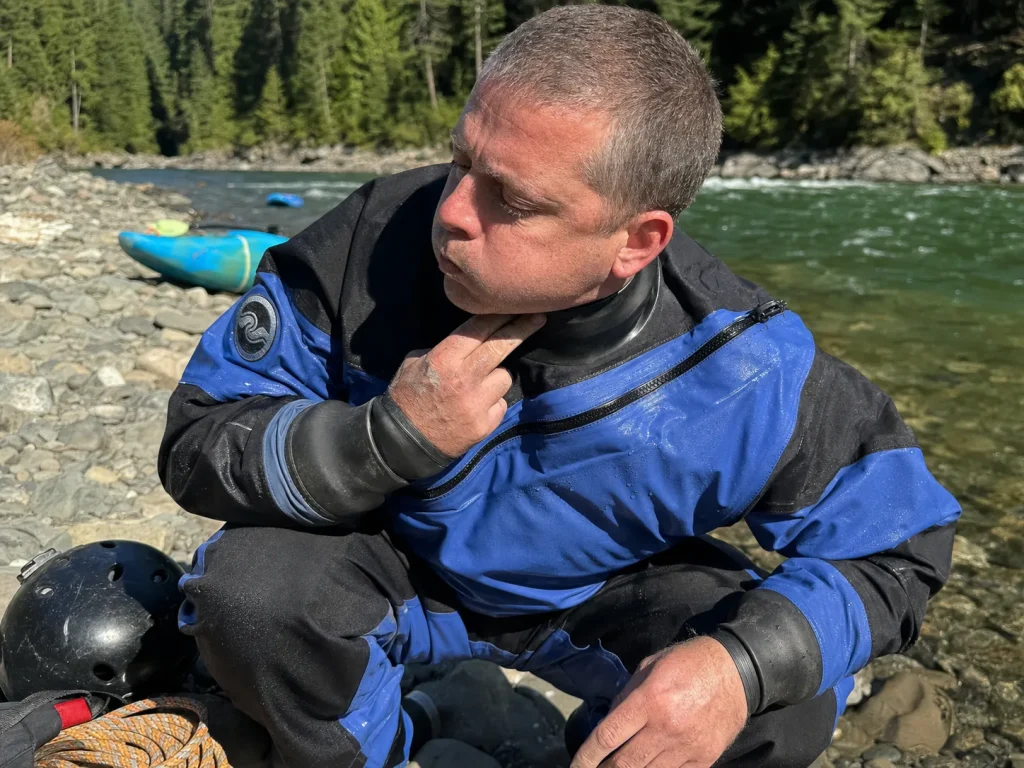 Rafter squatting to burp a drysuit at the water's edge, pushing trapped air out the neck gasket