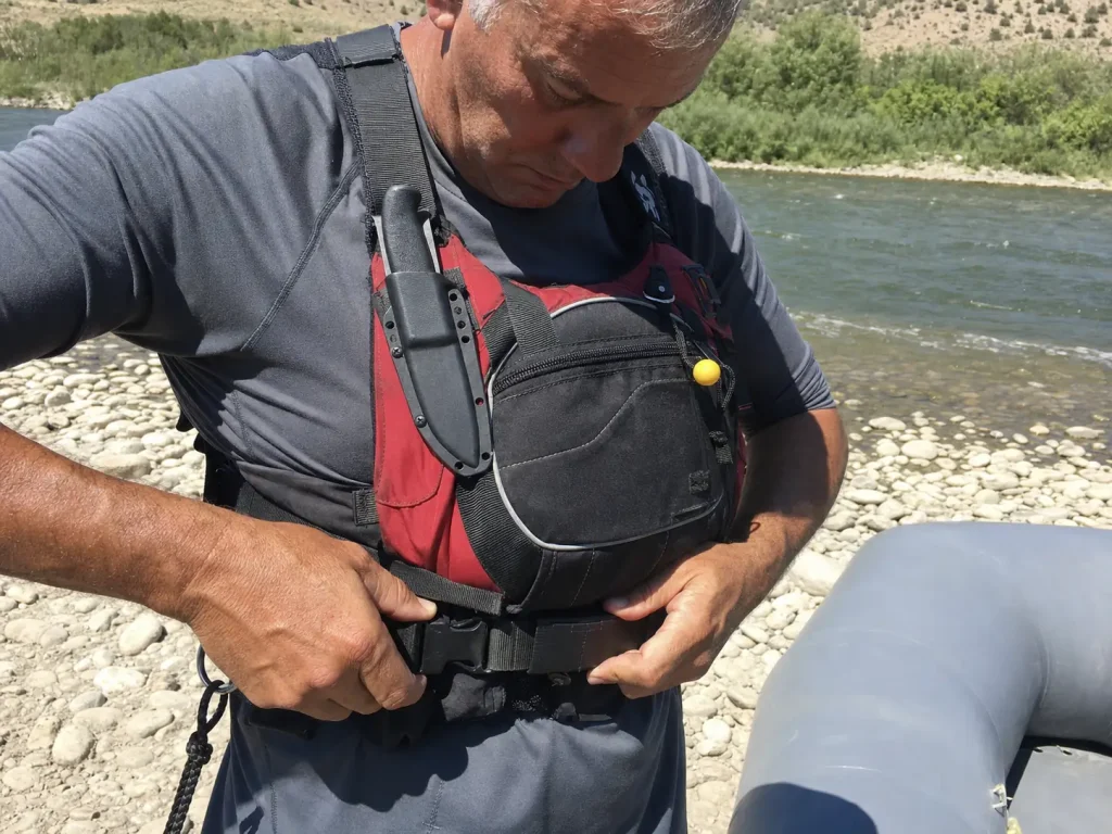 Hands finding the quick-release tab on a rescue PFD harness belt, the riskiest part of a river rescue kit
