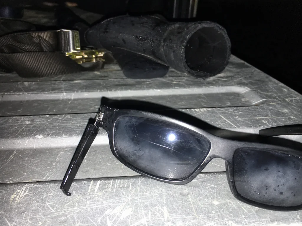 Beat-up cheap sunglasses with a cracked temple and fogged lens on a truck tailgate at dusk