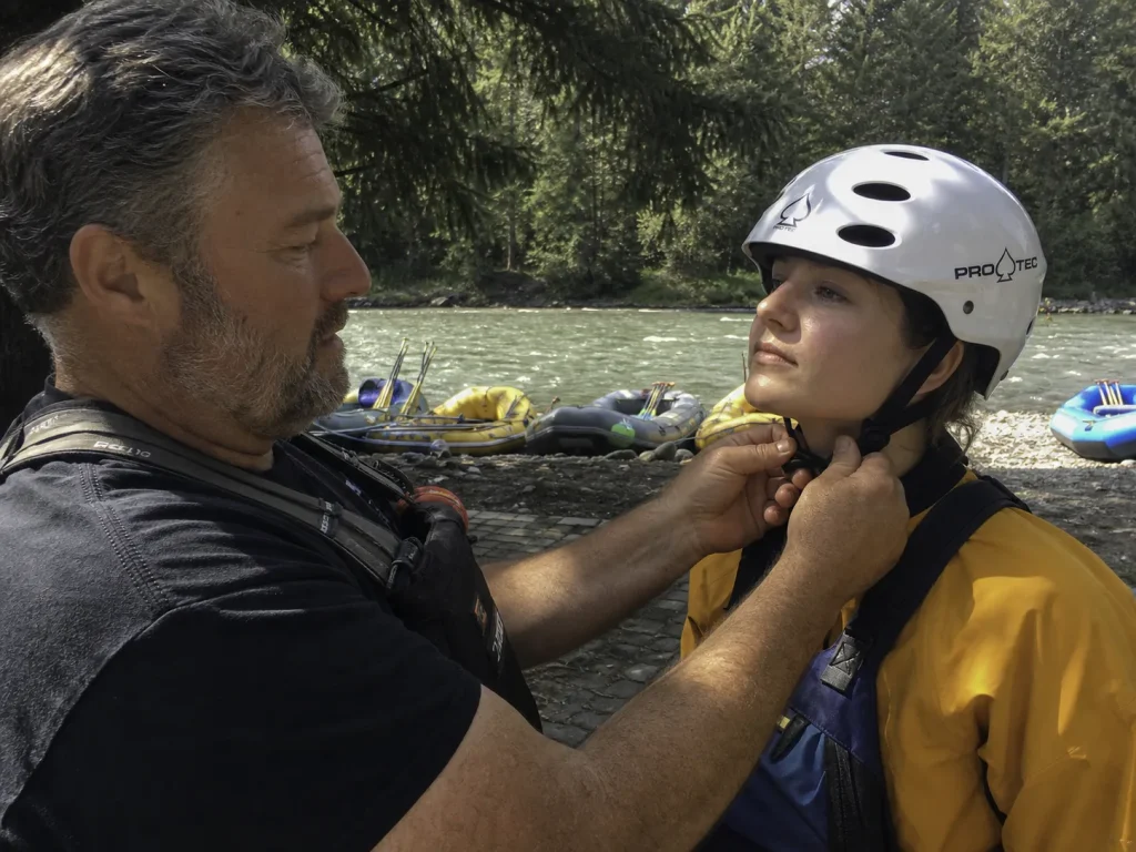 Guide checking a rafter's helmet strap tension by hand at the put-in before launching
