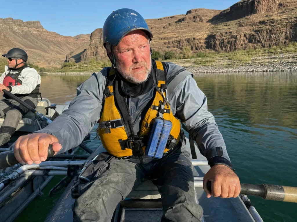 Rafter rowing an oar frame in a drysuit on a sunny stretch, sweating from the sustained effort