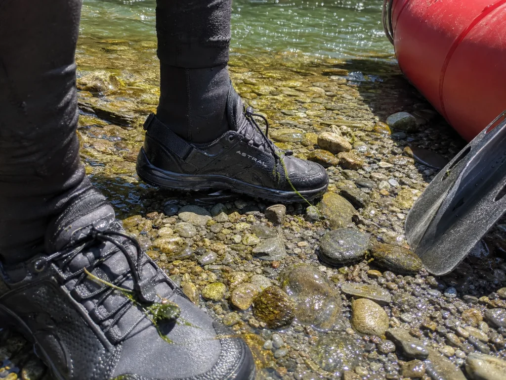 Neoprene socks in closed-toe Astral river shoes standing on wet gravel by a cold river