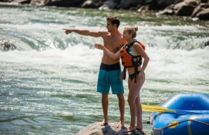 Best Water Shoes for Rafting: A Risk & Thermal Analysis A fit couple in swimwear and water shoes stand on a riverside rock, planning their route through whitewater rapids next to their raft.