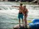 Best Water Shoes for Rafting: A Risk & Thermal Analysis A fit couple in swimwear and water shoes stand on a riverside rock, planning their route through whitewater rapids next to their raft.