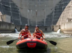 How Dams Affect Rivers: A Rafter’s Guide to Flow & Safety A man and woman in a whitewater raft on a sunny river, looking up at the massive concrete wall of a hydroelectric dam.