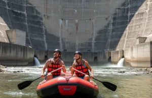 How Dams Affect Rivers: A Rafter’s Guide to Flow & Safety A man and woman in a whitewater raft on a sunny river, looking up at the massive concrete wall of a hydroelectric dam.