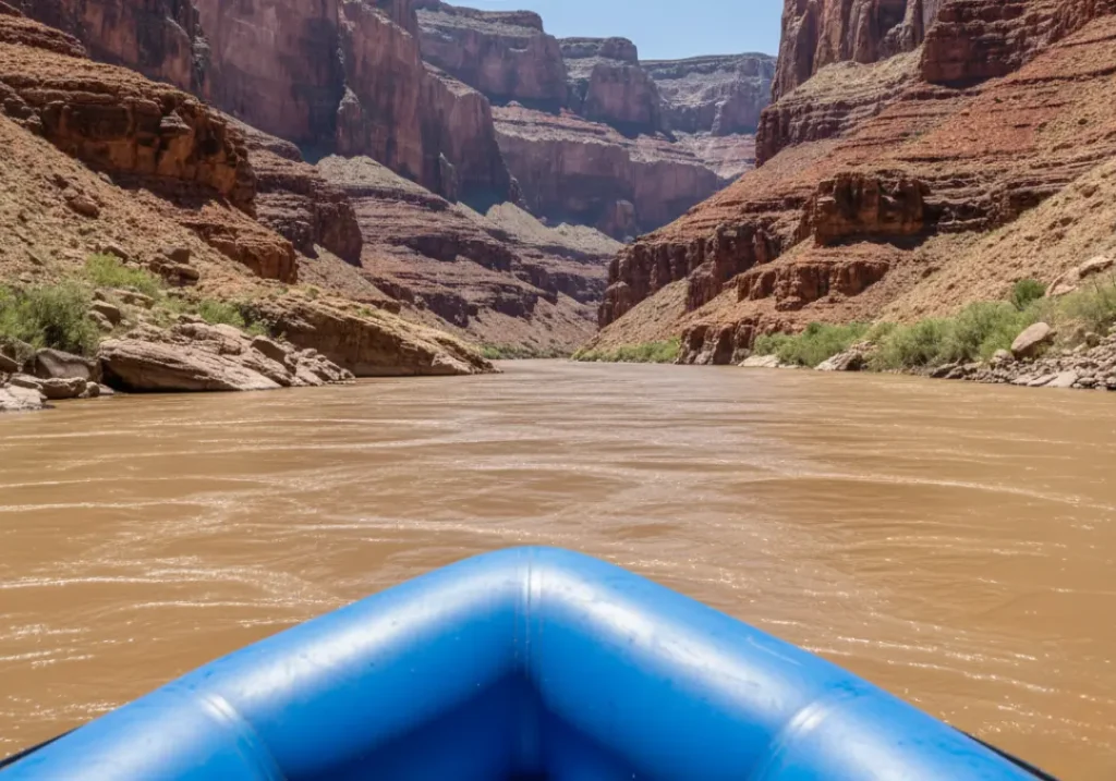 A scenic view down the Colorado River from a raft, showing the immense red rock walls of the Grand Canyon.