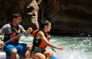 Guiding a Raft: From Basic Strokes to River Reading A full-body shot of a fit man and woman in their late 20s guiding a raft through whitewater rapids on a sunny day.