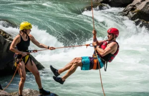 When to Use a Live Bait Rescue? A Complete Guide A male swiftwater rescuer in full gear is being lowered into a rapid while a female teammate belays him from the riverbank.