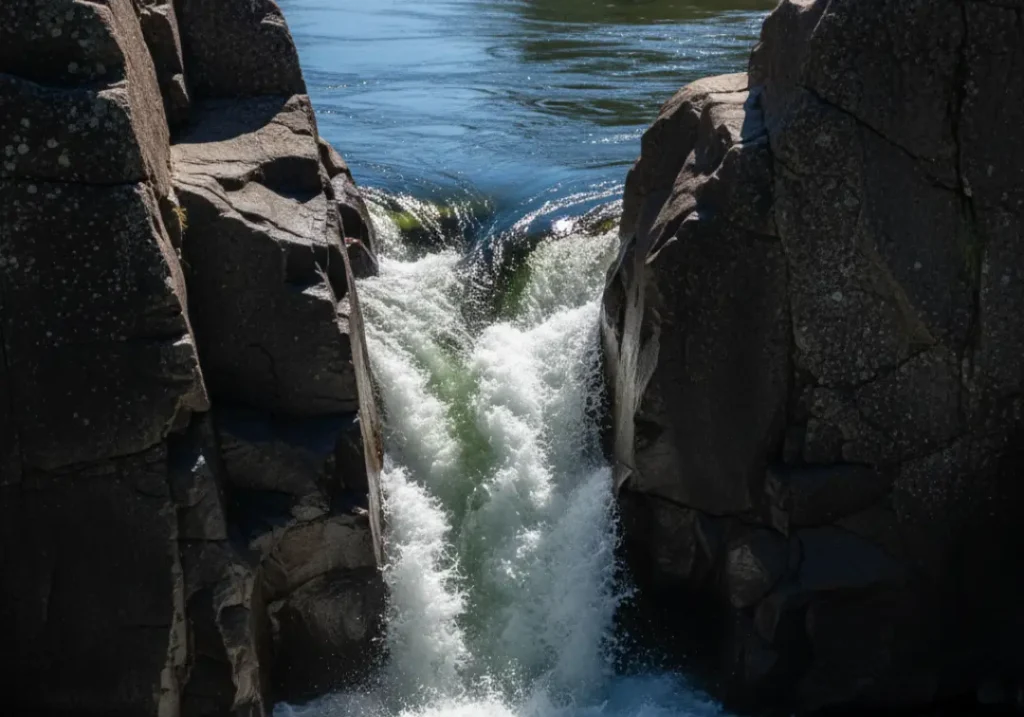 A river channel narrowing dramatically, transforming calm, smooth water into a chaotic, white rapid.