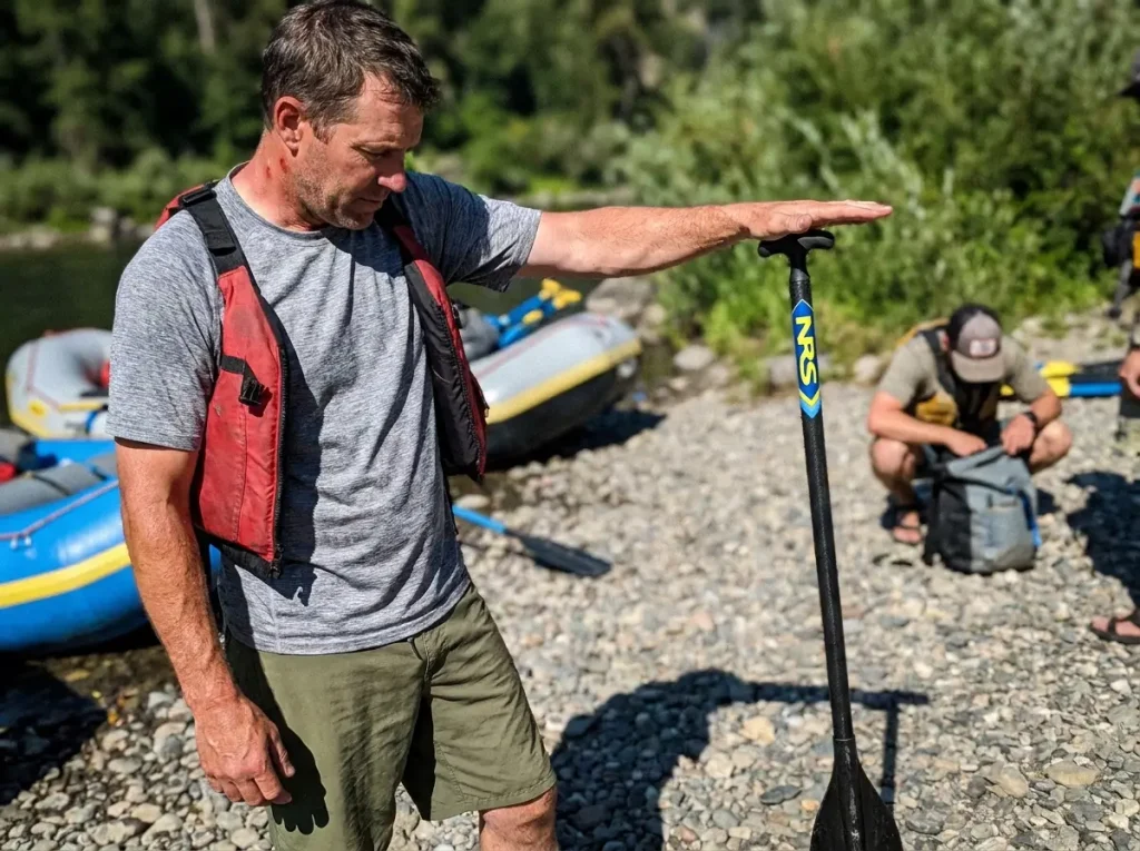 Paddler standing a raft paddle blade-down on gravel to measure length against mid-chest at the put-in
