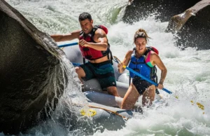 River Pinning Explained: Causes, Types & Rescue Protocol A full-body shot of a man and woman in a raft pinned sideways against a large rock in turbulent whitewater.