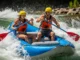 How to Surf a Raft: Mastering River Hydraulics & Flow Full-body shot of a fit man and woman in their late 20s, wearing swimwear, PFDs, and helmets, actively surfing a whitewater raft on a stationary river wave.