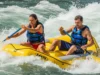 River Hazards Explained: A Rafter’s Field Manual A man and woman in a raft, both wearing PFDs, paddle through intense whitewater rapids on a sunny day.