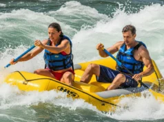River Hazards Explained: A Rafter’s Field Manual A man and woman in a raft, both wearing PFDs, paddle through intense whitewater rapids on a sunny day.
