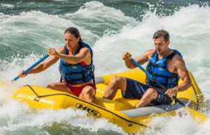 River Hazards Explained: A Rafter’s Field Manual A man and woman in a raft, both wearing PFDs, paddle through intense whitewater rapids on a sunny day.
