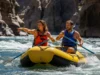 The Rafter’s CFS Impact Scale: Reading River Flows A full-body landscape shot of an athletic couple in their late 20s in a raft, studying the whitewater ahead on a sunny day.