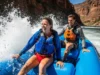 Outfitter-Tested Splash Jackets: A Definitive Guide A fit man and woman in their late 20s, wearing splash jackets and PFDs over swimsuits, paddle an inflatable raft through whitewater rapids on a sunny day.