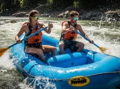 Best Rafting Sunglasses: Tested for Impact & Retention A fit couple in their late 20s wearing life jackets and sunglasses paddle a raft through a sunny whitewater rapid.