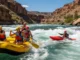 Rafting vs Kayaking: A Rafter’s Technical Breakdown A full-body, wide-angle shot of a group of four people in a raft and a solo kayaker navigating a sunny whitewater rapid.