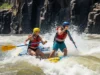 Rafting Zambezi River: A Technical Rapids Analysis (2025) A fit man and woman paddle furiously in a raft, crashing through a huge whitewater rapid on the Zambezi River, framed by the towering walls of the Batoka Gorge.