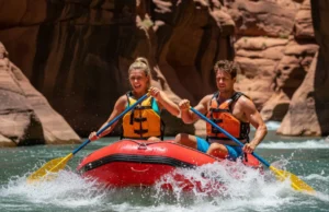 The Science of Rapids: How They Form & How to Read Them A full-body shot of a young man and woman in a raft navigating a sunny whitewater rapid in a deep canyon.