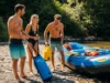 Mastering River Etiquette: A Code for Whitewater A full-body shot of two men and a woman in swimwear, laughing on a sunny riverbank as they prepare their raft for a whitewater trip.