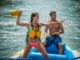 Best River Rescue Throw Bags: Rated for Raft Rescue A fit woman in a bikini and life jacket throws a rescue bag from a raft on a sunny day, with a man assisting her.
