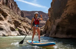River SUP Guide: A Progressive & Responsible Framework A full-body shot of a woman in a swimsuit, PFD, and helmet stand-up paddleboarding on a sunny river.