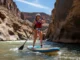 River SUP Guide: A Progressive & Responsible Framework A full-body shot of a woman in a swimsuit, PFD, and helmet stand-up paddleboarding on a sunny river.