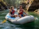 Mastering the Rogue: A Tactical River Rafting Guide A fit young couple in a raft navigate a sunny, tree-lined canyon on the Rogue River.