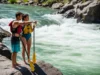 How to Scout a Rapid: The Guide’s Field Blueprint A fit couple in their late 20s, wearing PFDs and swimwear, stand on a rock scouting a whitewater rapid from the riverbank.