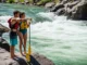 How to Scout a Rapid: The Guide’s Field Blueprint A fit couple in their late 20s, wearing PFDs and swimwear, stand on a rock scouting a whitewater rapid from the riverbank.