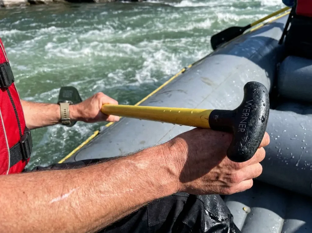 Close-up of hands gripping a raft paddle T-grip braced over the tube showing too-long paddle leverage