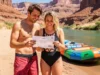 An Analysis of USA River Permits: Odds, Strategy & Skills A man and woman in swimwear stand on a riverbank in a canyon, smiling as they hold a river permit together next to their raft.