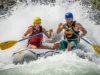 Cold Shock & Breathing in Whitewater: A Rafter’s Protocol A full-body shot of an athletic couple in their late 20s navigating a whitewater rapid on a raft, getting splashed by a large wave.