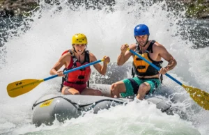 Cold Shock & Breathing in Whitewater: A Rafter’s Protocol A full-body shot of an athletic couple in their late 20s navigating a whitewater rapid on a raft, getting splashed by a large wave.