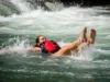 Whitewater Swim Position: Defensive vs. Aggressive Guide A full-body shot of a young woman in a one-piece swimsuit and PFD floating on her back in the defensive swim position down a whitewater rapid.