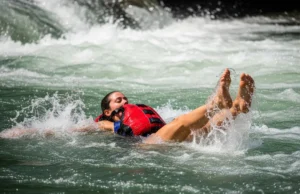 Whitewater Swim Position: Defensive vs. Aggressive Guide A full-body shot of a young woman in a one-piece swimsuit and PFD floating on her back in the defensive swim position down a whitewater rapid.