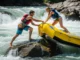 Raft Wrap-Risk Score: A Guide to Prevention & Recovery A full-body shot of a young man and woman in swimwear and PFDs working together to unpin their raft from a large boulder in a whitewater rapid.