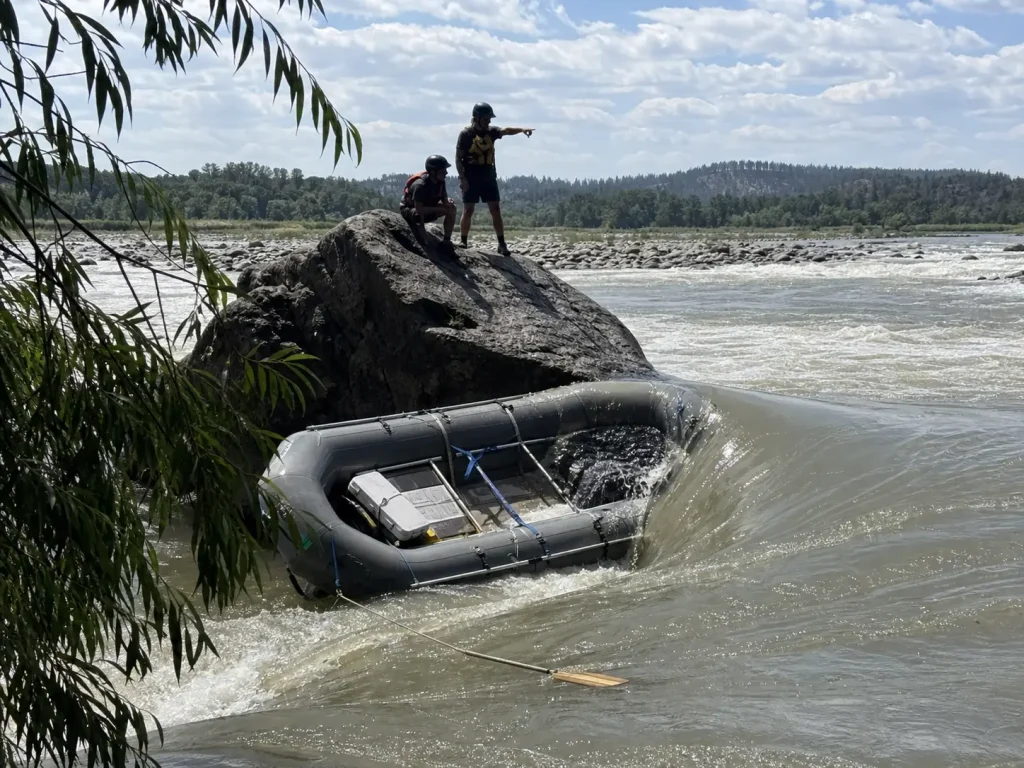 A loaded raft pinned against a midstream boulder showing why raft pin kit loads exceed kayak loads