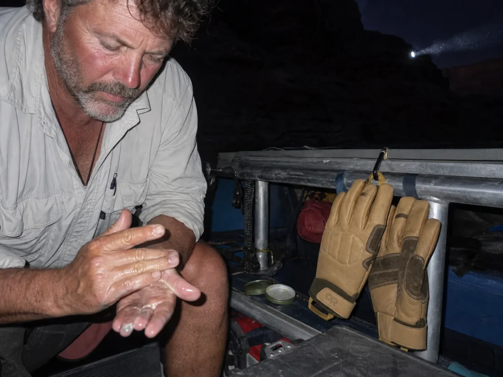Leather rafting gloves drying on a raft frame at camp with hands rubbing in salve at dusk