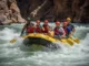 The Definitive Rafting Carnage Guide (Safety & Skills) A group of rafters led by a skilled guide successfully maneuvering their raft through a large and challenging whitewater rapid in a sunny canyon.