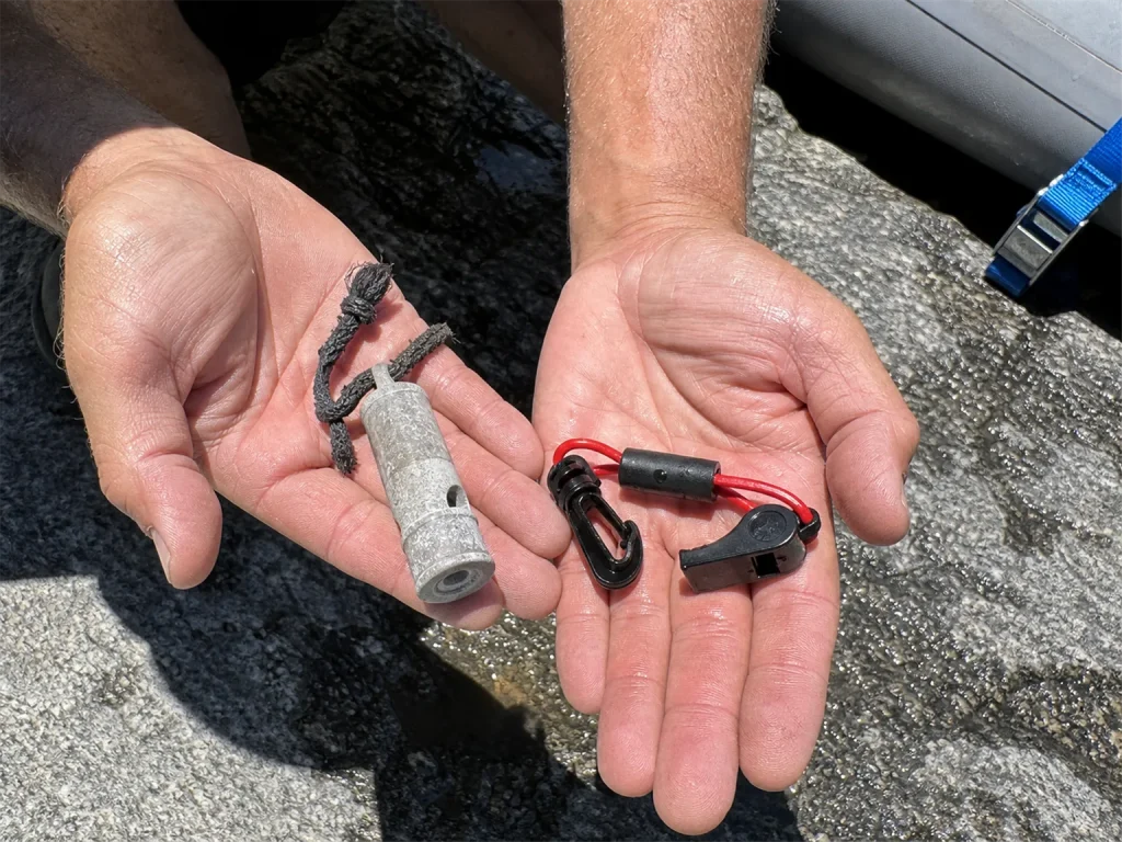 Hands holding a pealess paddle whistle and an old pea whistle side by side on a wet rock